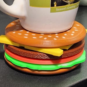 May include: A brown and white patterned mug sits on top of a multi-layered, brown, yellow, red, and green burger-shaped coaster. The mug has the text "Season's Greetings" on the side.