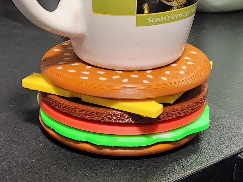 May include: A brown and white patterned mug sits on top of a multi-layered, brown, yellow, red, and green burger-shaped coaster. The mug has the text "Season's Greetings" on the side.