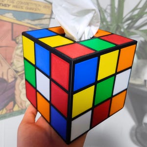 May include: A tissue box shaped like a Rubik's Cube with red, blue, yellow, green, orange, and white squares. The tissue box is made of plastic and has a black frame.