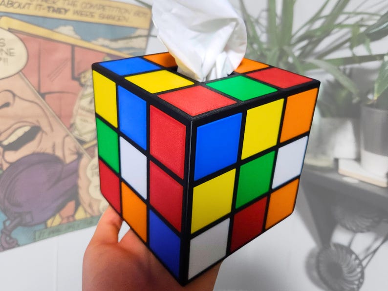 May include: A tissue box shaped like a Rubik's Cube with red, blue, yellow, green, orange, and white squares. The tissue box is made of plastic and has a black frame.