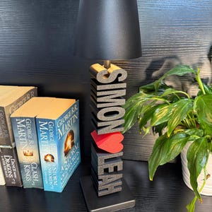 May include: A black lamp with a black shade sits on a wooden base with the names "Simon" and "Leah" spelled out in large black letters. A red heart is between the names. The lamp is on a black surface with a stack of books and a green plant in a white pot.