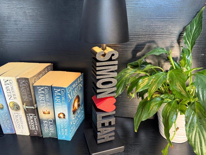 May include: A black lamp with a black shade sits on a wooden base with the names "Simon" and "Leah" spelled out in large black letters. A red heart is between the names. The lamp is on a black surface with a stack of books and a green plant in a white pot.