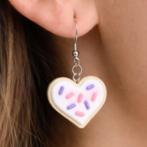 May include: A heart-shaped cookie earring with white icing and colorful sprinkles. The earring is made of a light beige material with a silver-colored hook and chain. The sprinkles are pink and purple.