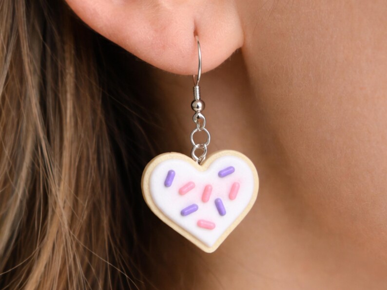 May include: A heart-shaped cookie earring with white icing and colorful sprinkles. The earring is made of a light beige material with a silver-colored hook and chain. The sprinkles are pink and purple.