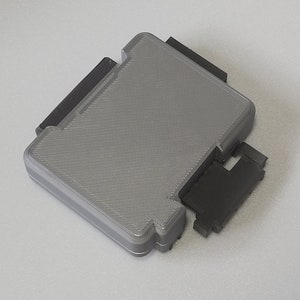 May include: A gray plastic case with a textured surface and black plastic hinges. The case is closed and the hinges are visible.