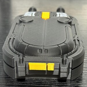 May include: A black and yellow rectangular device with a textured surface. The device has a rounded top and bottom with yellow accents. The design includes two small, round protrusions on top.
