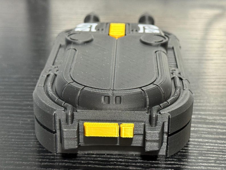 May include: A black and yellow rectangular device with a textured surface. The device has a rounded top and bottom with yellow accents. The design includes two small, round protrusions on top.