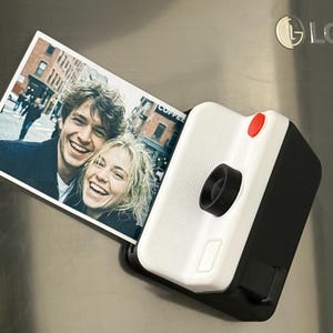 May include: A white and black camera-shaped fridge magnet displaying a photograph of two smiling people. The magnet has a red button and a black lens. The photograph shows a couple smiling in front of a city backdrop.