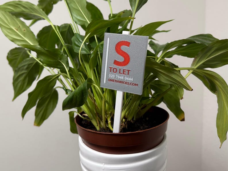 May include: A potted plant with large green leaves. A gray sign with the red letter "S" and the words "TO LET" is placed in the plant. The plant is in a brown pot, which sits on a white, ribbed planter.