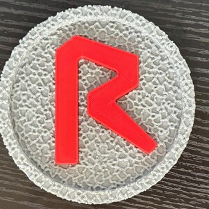 May include: A round, gray coaster with a textured surface and a bright red letter "R" in the center. The coaster is likely made of a durable material and is designed to protect surfaces from drink condensation.