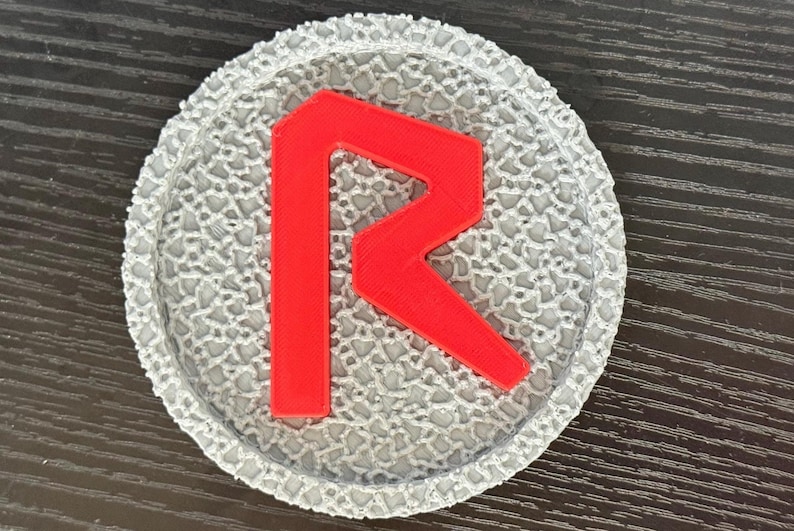May include: A round, gray coaster with a textured surface and a bright red letter "R" in the center. The coaster is likely made of a durable material and is designed to protect surfaces from drink condensation.