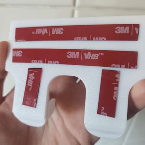 May include: White plastic wall mount with four red 3M VHB adhesive strips. The mount has a hook shape with a hole in the center.