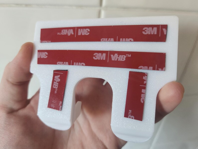 May include: White plastic wall mount with four red 3M VHB adhesive strips. The mount has a hook shape with a hole in the center.