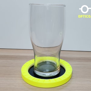 May include: A yellow circular coaster with a black center and a glass on top. The coaster has the text "OPTICOASTER" printed on it.