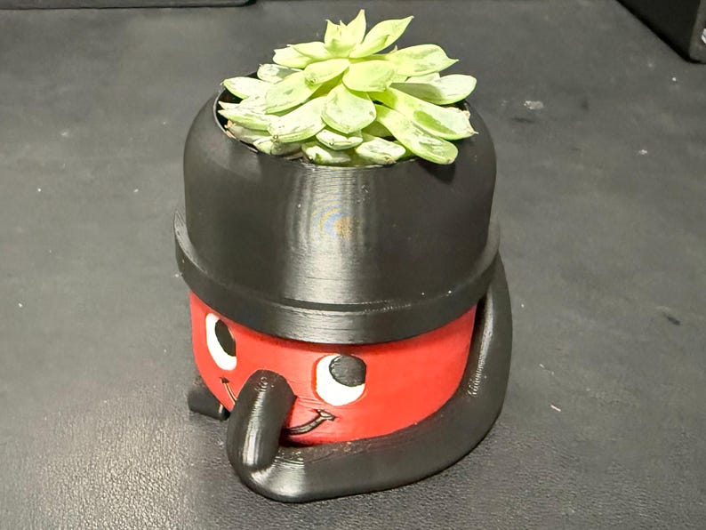 May include: A 3D-printed planter shaped like a cartoon vacuum cleaner, featuring a red body, black accents, and a smiling face. The planter holds a succulent with green and white leaves. A unique and playful home decor item.