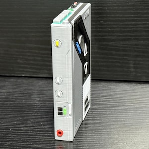 May include: A silver and black portable audio player with a retro design. It features buttons, switches, and a headphone jack. The device has a rectangular shape with a textured surface and a small screen.