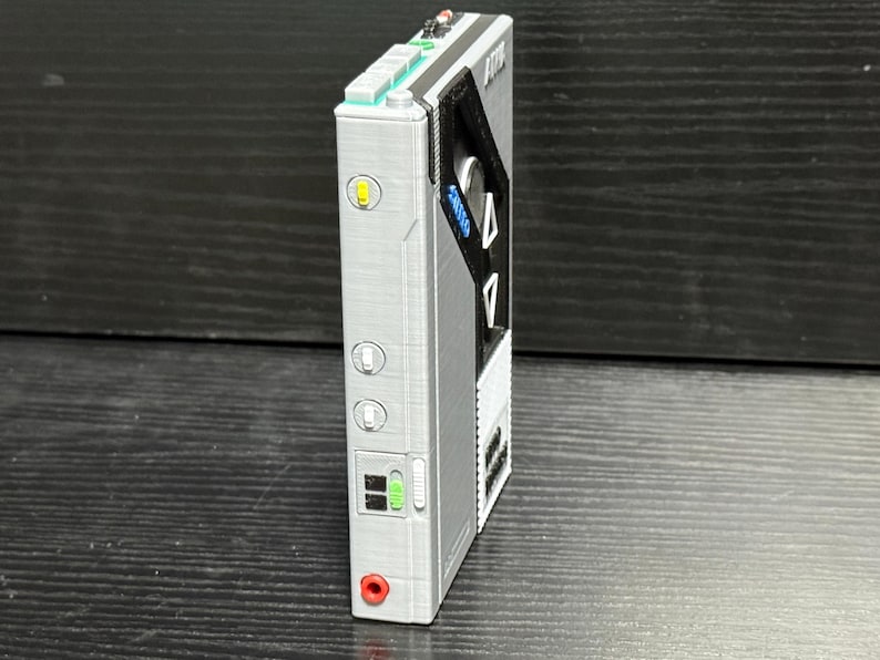 May include: A silver and black portable audio player with a retro design. It features buttons, switches, and a headphone jack. The device has a rectangular shape with a textured surface and a small screen.