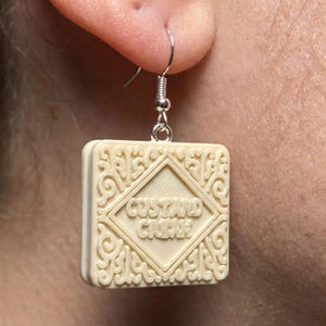 Puede incluir: Pendiente individual de galleta Custard Cream de color marfil con gancho plateado. El pendiente cuadrado presenta un centro en forma de diamante con las palabras "Custard Cream" y adornos decorativos. Un accesorio &uacute;nico con tem&aacute;tica de comida.