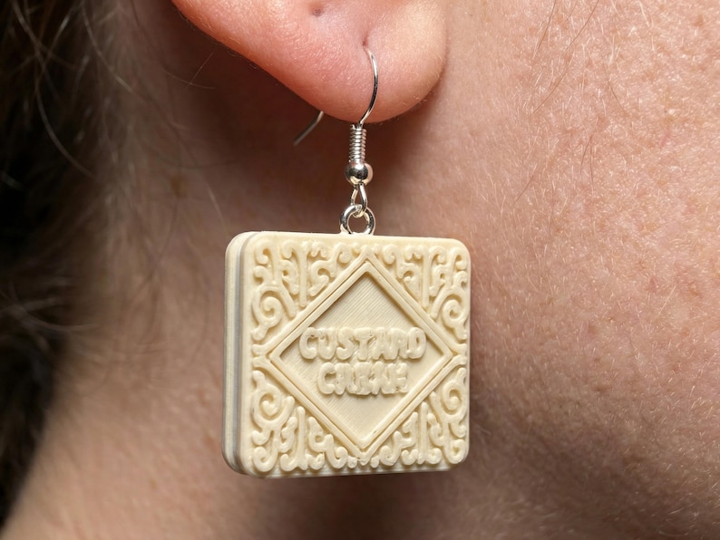 Puede incluir: Pendiente individual de galleta Custard Cream de color marfil con gancho plateado. El pendiente cuadrado presenta un centro en forma de diamante con las palabras "Custard Cream" y adornos decorativos. Un accesorio &uacute;nico con tem&aacute;tica de comida.