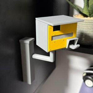 K&ouml;nnte beinhalten: Ein 3D-gedrucktes Modell eines Radarblitzers, mit grauem Geh&auml;use und gelber Frontplatte. Es hat ein Dach und einen Montagearm zur Wandbefestigung. Das Modell ist eine Miniaturreplik.