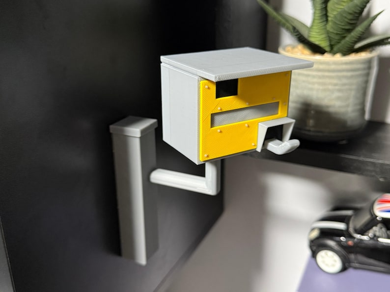 K&ouml;nnte beinhalten: Ein 3D-gedrucktes Modell eines Radarblitzers, mit grauem Geh&auml;use und gelber Frontplatte. Es hat ein Dach und einen Montagearm zur Wandbefestigung. Das Modell ist eine Miniaturreplik.