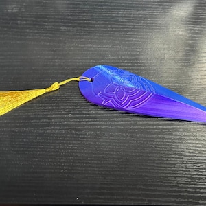 May include: A blue and purple bookmark with a gold tassel. The bookmark has a floral design and a pointed end. The tassel is attached to the top of the bookmark with a small loop. The bookmark is lying on a dark surface.