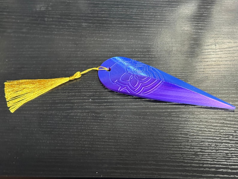 May include: A blue and purple bookmark with a gold tassel. The bookmark has a floral design and a pointed end. The tassel is attached to the top of the bookmark with a small loop. The bookmark is lying on a dark surface.