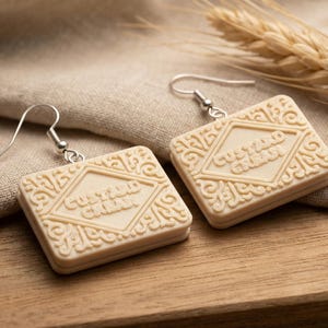 Puede incluir: Un par de pendientes de galleta Custard Cream de color crema con ganchos plateados. Los pendientes presentan las palabras "Custard Cream" en un dise&ntilde;o en forma de diamante, rodeado de patrones decorativos. Se exhiben sobre tela beige y madera.