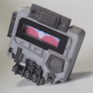 May include: A gray 3D printed device with a small screen displaying a red, white, and blue flag with a snowflake design. The device has several buttons and a textured surface.