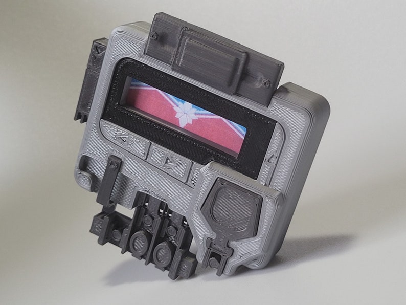 May include: A gray 3D printed device with a small screen displaying a red, white, and blue flag with a snowflake design. The device has several buttons and a textured surface.