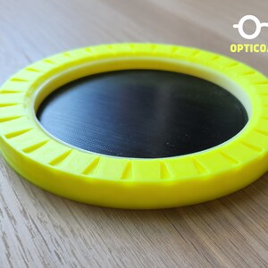 May include: A yellow circular coaster with a black center and a white ring around the edge. The coaster has a textured design around the edge. The text "OPTICOASTER" is printed on the coaster.