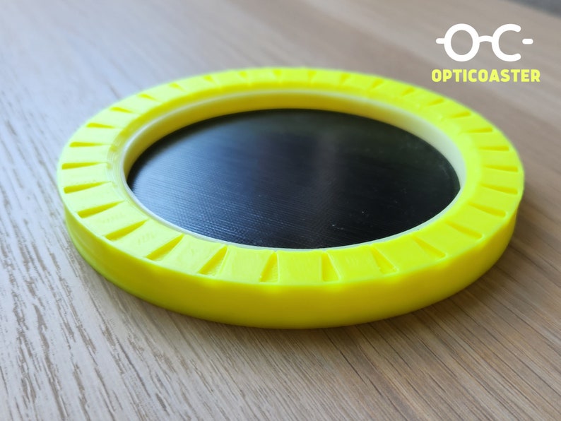 May include: A yellow circular coaster with a black center and a white ring around the edge. The coaster has a textured design around the edge. The text "OPTICOASTER" is printed on the coaster.