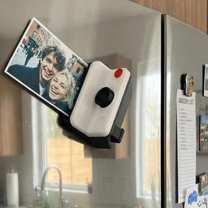 May include: A white and black camera-shaped fridge magnet holding a photograph of two smiling people. The magnet features a red button and a black lens. Other magnets and a grocery list are also on the refrigerator.