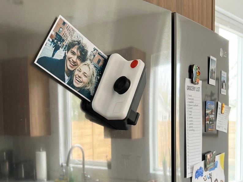 May include: A white and black camera-shaped fridge magnet holding a photograph of two smiling people. The magnet features a red button and a black lens. Other magnets and a grocery list are also on the refrigerator.