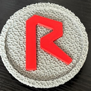 May include: A gray, textured coaster with a red letter "R" in the center. The coaster has a circular shape with a detailed, patterned border. The red letter stands out against the gray background.