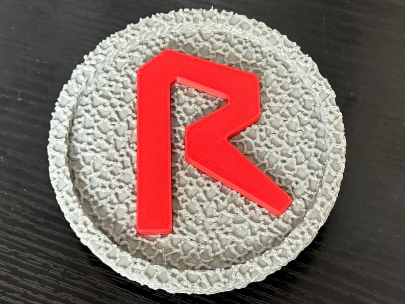 May include: A gray, textured coaster with a red letter "R" in the center. The coaster has a circular shape with a detailed, patterned border. The red letter stands out against the gray background.