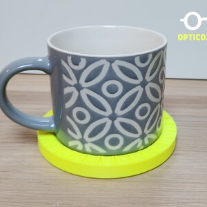May include: A gray ceramic mug with a white geometric pattern sits on a bright yellow circular coaster. The coaster has a textured surface and a raised edge.