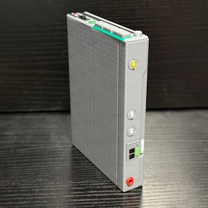 May include: A gray rectangular electronic device with a series of buttons, lights, and ports. The device has a green and white top section with small buttons. A yellow light is visible on the side, along with two circular buttons and a red port.