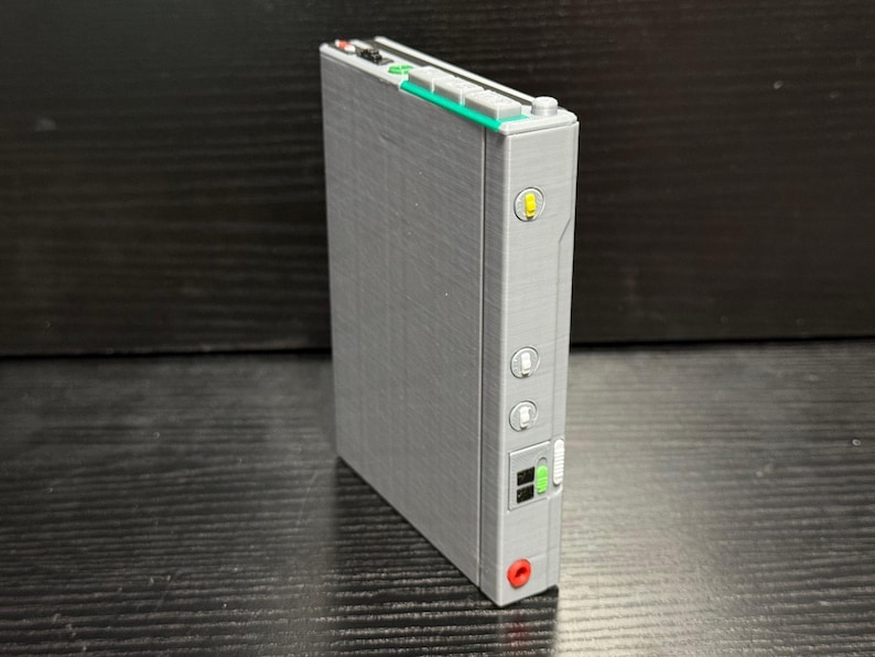 May include: A gray rectangular electronic device with a series of buttons, lights, and ports. The device has a green and white top section with small buttons. A yellow light is visible on the side, along with two circular buttons and a red port.