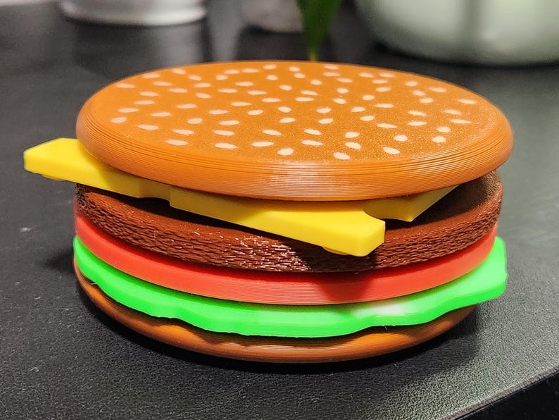 May include: A toy hamburger made of plastic with a brown bun, yellow cheese, red tomato, green lettuce, and a brown patty.