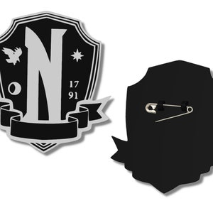 May include: Black and silver shield-shaped pin with a large letter N, a small bird, a star, and the numbers 17 and 91. The back of the pin has a safety pin.