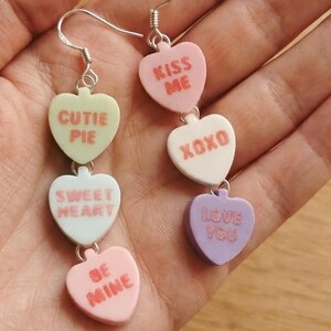 May include: Dangling earrings featuring heart-shaped candy charms in pastel colors. Each charm has a different message, including "Cutie Pie," "Sweet Heart," "Be Mine," "Kiss Me," "XOXO," and "Love You." The earrings are attached to silver-colored fish hook ear wires.