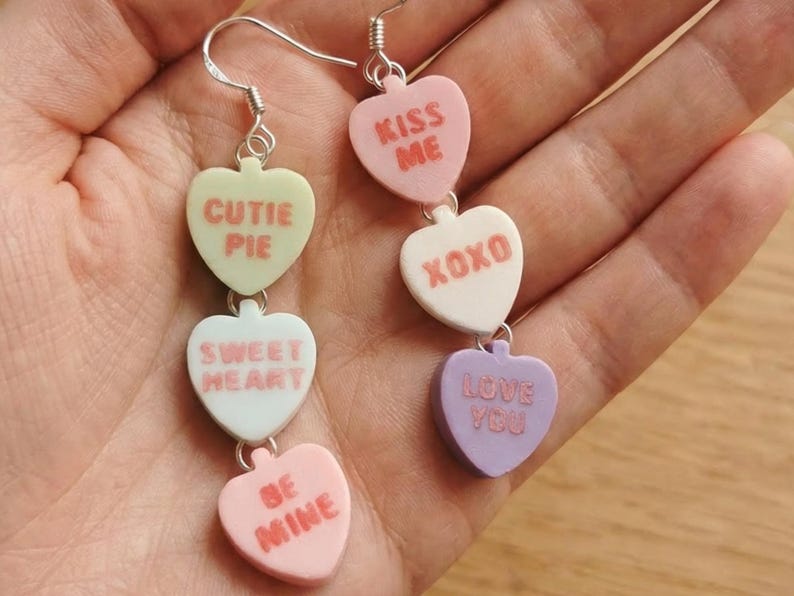 May include: Dangling earrings featuring heart-shaped candy charms in pastel colors. Each charm has a different message, including "Cutie Pie," "Sweet Heart," "Be Mine," "Kiss Me," "XOXO," and "Love You." The earrings are attached to silver-colored fish hook ear wires.