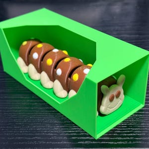 May include: A 3D-printed toy caterpillar in a bright green box. The caterpillar is brown with yellow and white accents. The box is a vibrant green, and the caterpillar's head is visible at the end.