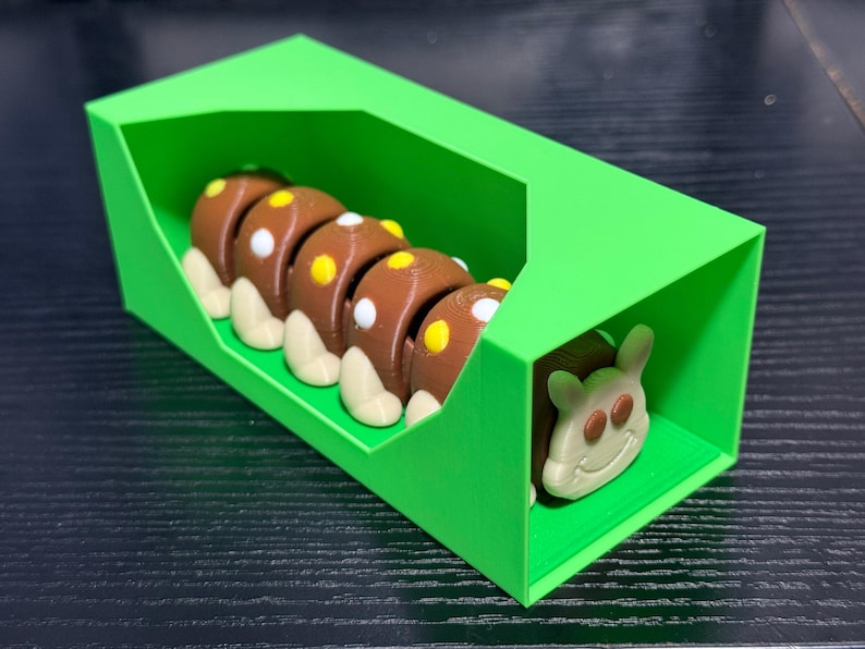 May include: A 3D-printed toy caterpillar in a bright green box. The caterpillar is brown with yellow and white accents. The box is a vibrant green, and the caterpillar's head is visible at the end.