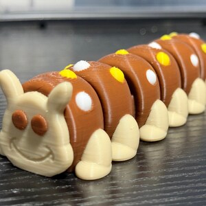 May include: A 3D-printed caterpillar toy in brown and cream colors. The caterpillar has a smiling face and segmented body with white and yellow accents. The toy is designed to be a fun and engaging item for children.