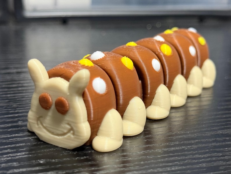 May include: A 3D-printed caterpillar toy in brown and cream colors. The caterpillar has a smiling face and segmented body with white and yellow accents. The toy is designed to be a fun and engaging item for children.