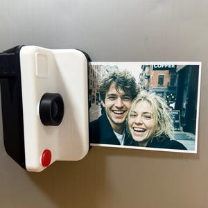 May include: A black and white instant camera-shaped magnet holding a colour photograph of a smiling couple. The camera has a red button and a black lens. The photograph shows a couple smiling in front of a building.