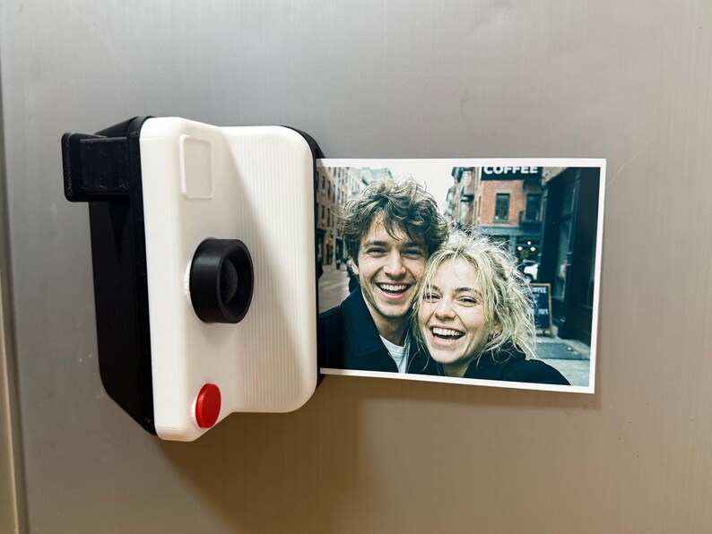 May include: A black and white instant camera-shaped magnet holding a colour photograph of a smiling couple. The camera has a red button and a black lens. The photograph shows a couple smiling in front of a building.