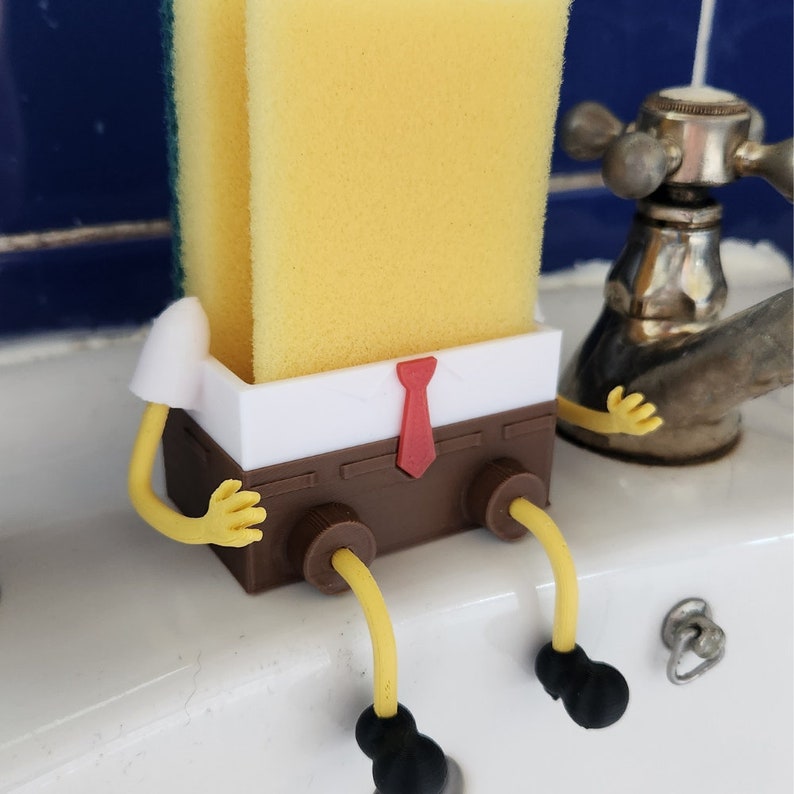 Puede incluir: Una esponja amarilla se encuentra en un soporte marr&oacute;n y blanco dise&ntilde;ado como un personaje de dibujos animados. El soporte tiene brazos y piernas amarillas, una corbata roja y zapatos negros. La esponja est&aacute; sobre una superficie blanca.